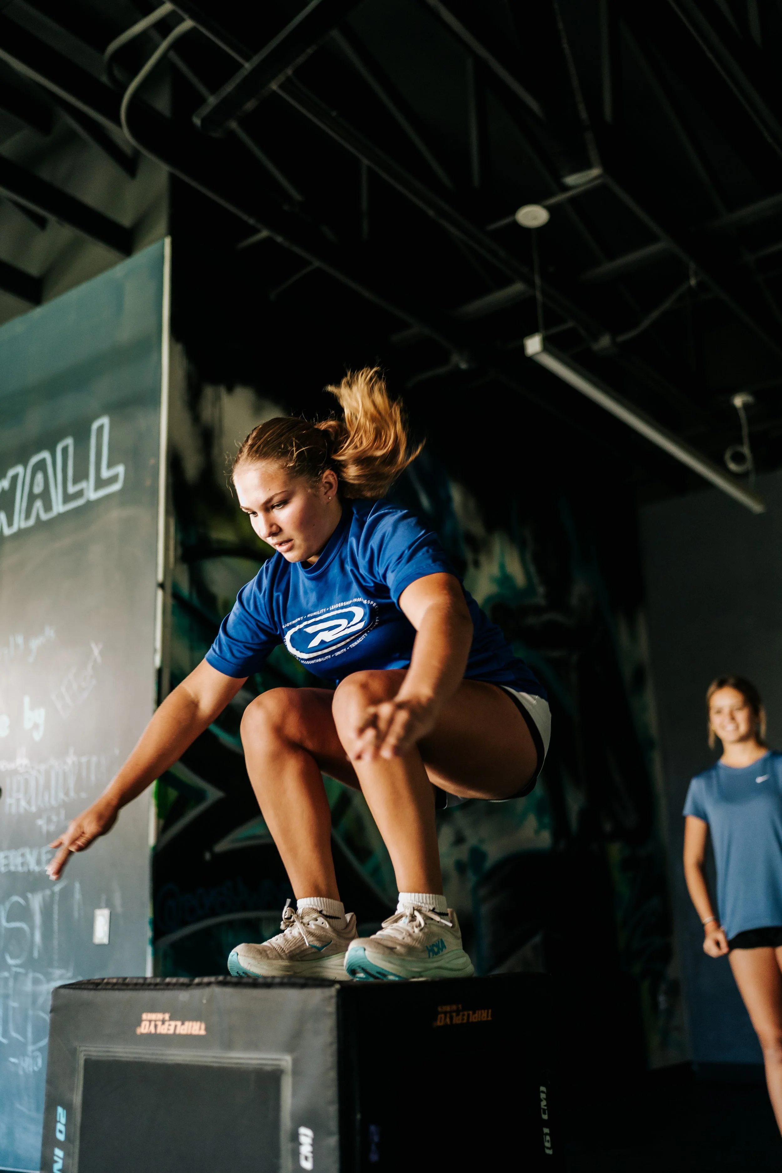 A young female athlete performs a box jump during a Breaking Lines strength and conditioning session as another athlete watches.