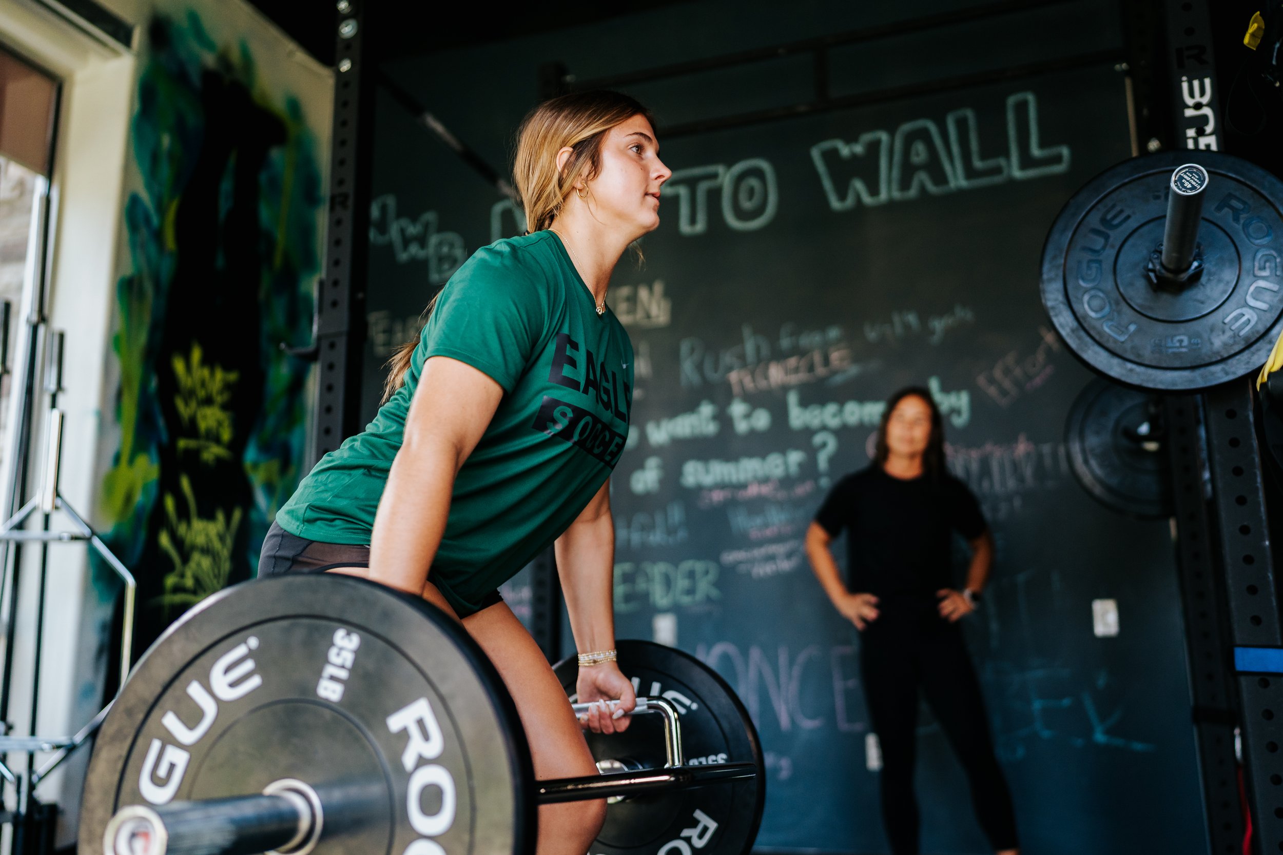 A young female athlete performs a trap bar deadlift during a Breaking Lines strength training session as founder and coach Ali Ohashi observes in the background.