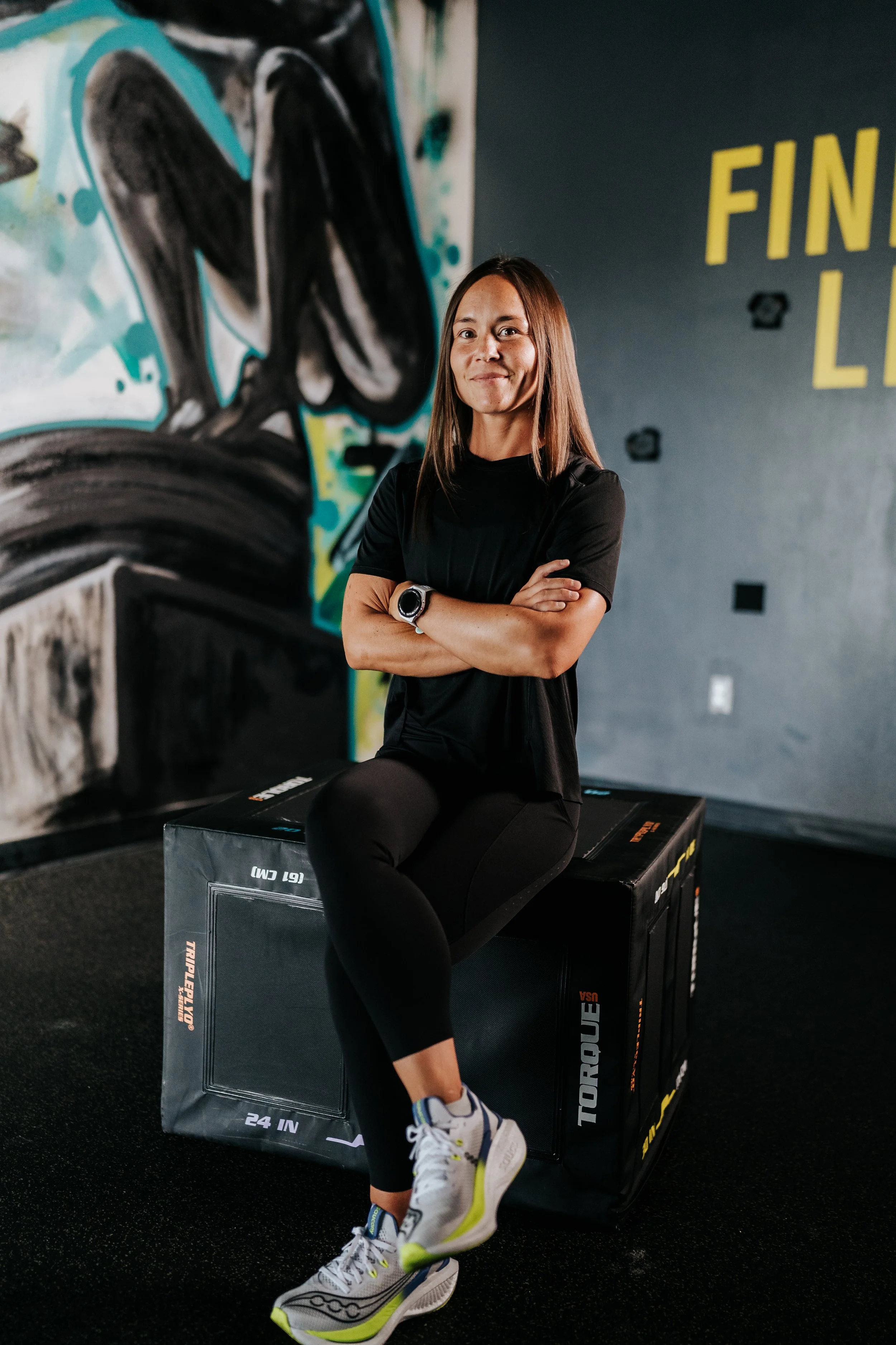 Breaking Lines founder and strength and conditioning coach Ali Ohashi sits on a plyometric jump box with her arms crossed inside a training gym in Boise, Idaho.