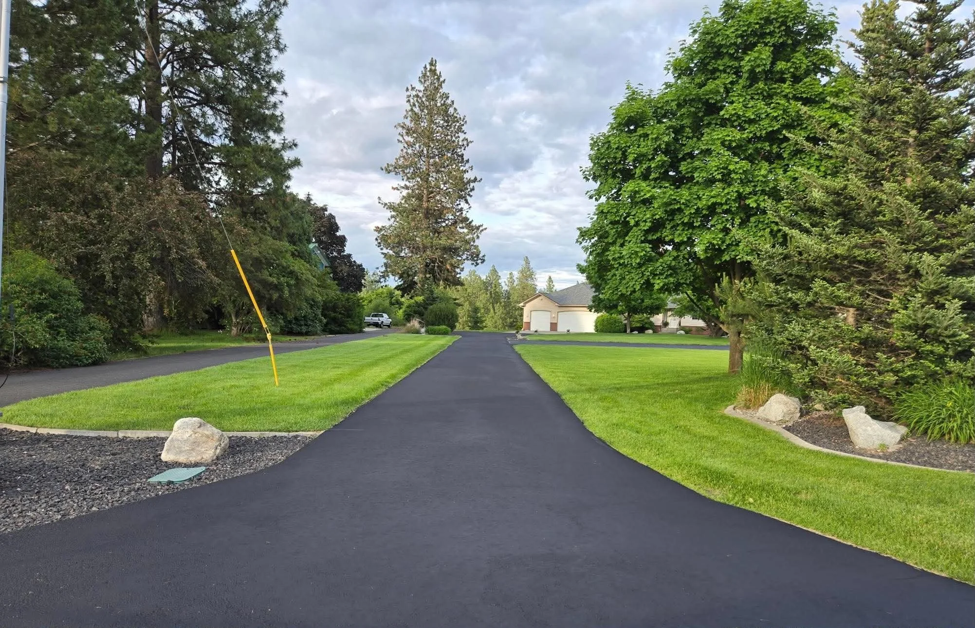 A newly paved asphalt driveway in a residential neighborhood on a cloudy day. The driveway is flanked by well-maintained green grass and landscaped areas with rocks. Tall trees and houses are visible in the background.