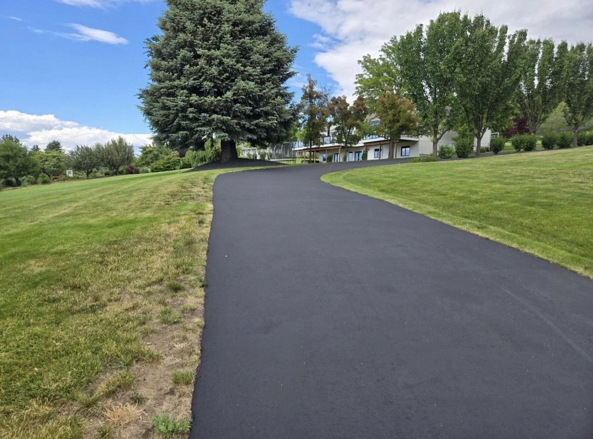 A freshly paved black asphalt pathway winding through a green park or yard with grass, trees, and a house in the background under a partly cloudy sky.