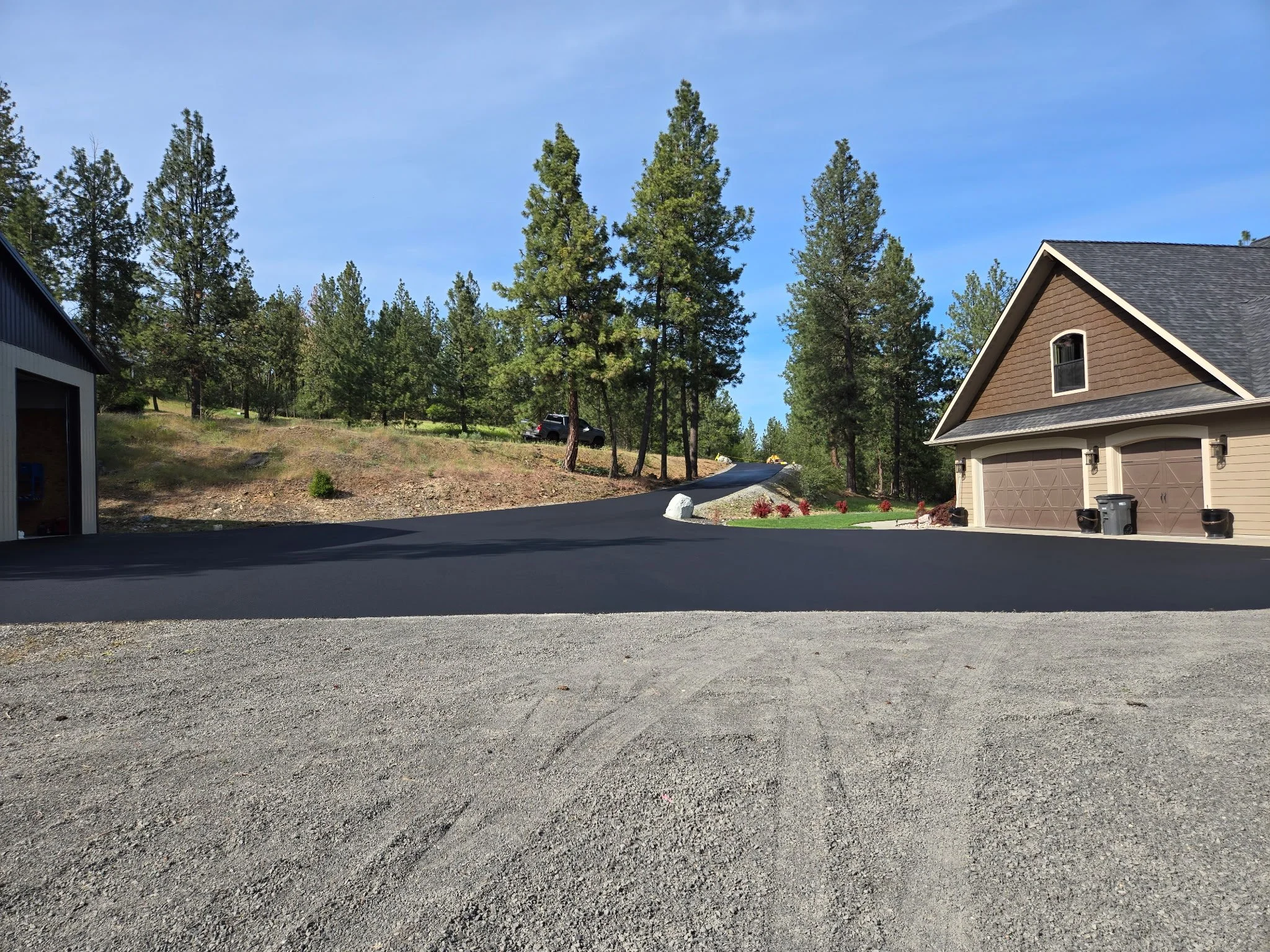A residential neighborhood with tall pine trees, a driveway, a house with a brown garage door, and some landscaping with flowers. The sky is clear and blue.