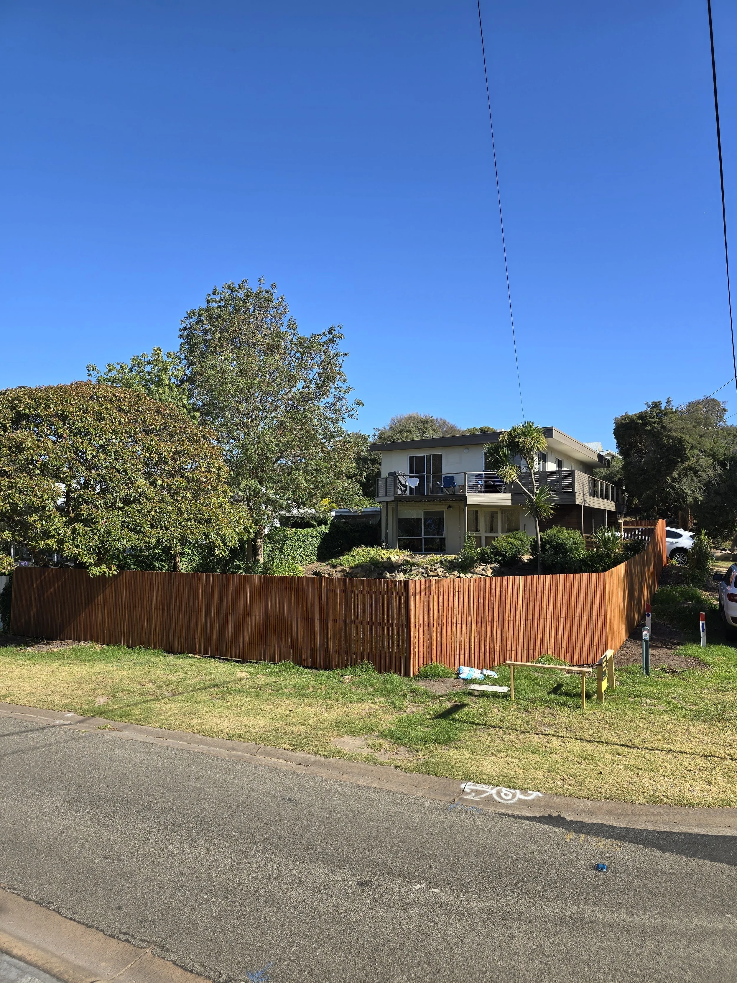 A two-story house with a balcony, surrounded by trees and a wooden fence, located behind a curved road.
