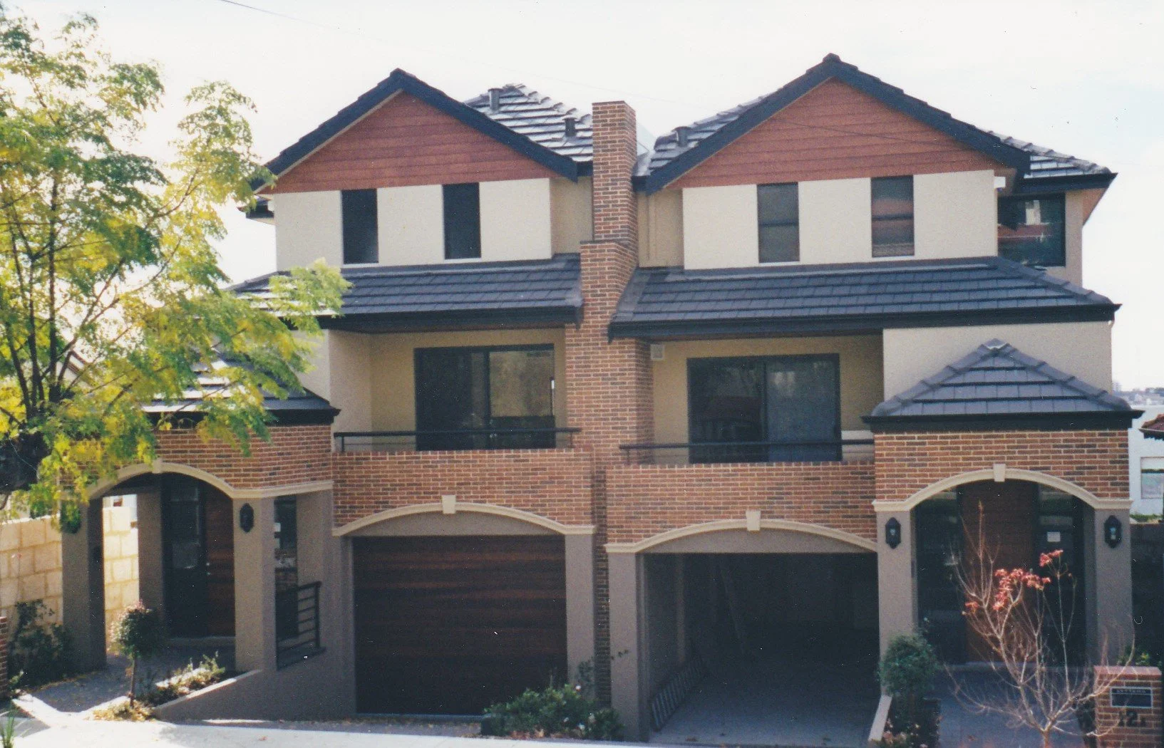 a multi-story house with brick and stucco exterior, black roof, and multiple balconies, surrounded by trees and plants