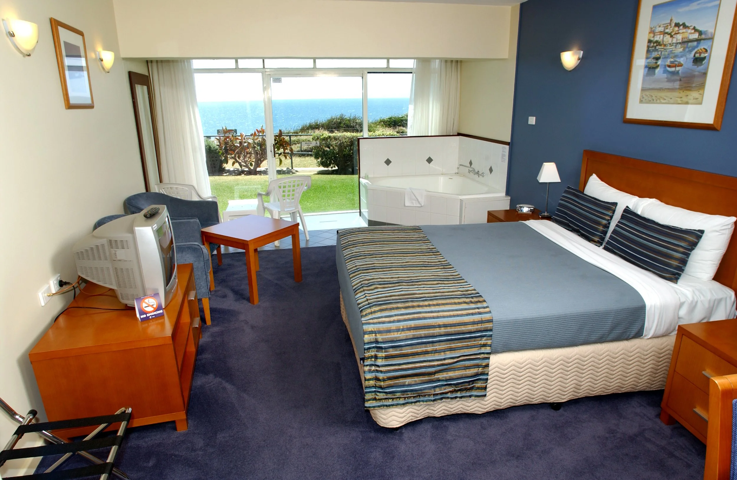 Hotel room with a large window view of the ocean, a bed with striped bedding, a small table, chairs, a bathtub near the window, a TV on a wooden stand, and wall paintings.