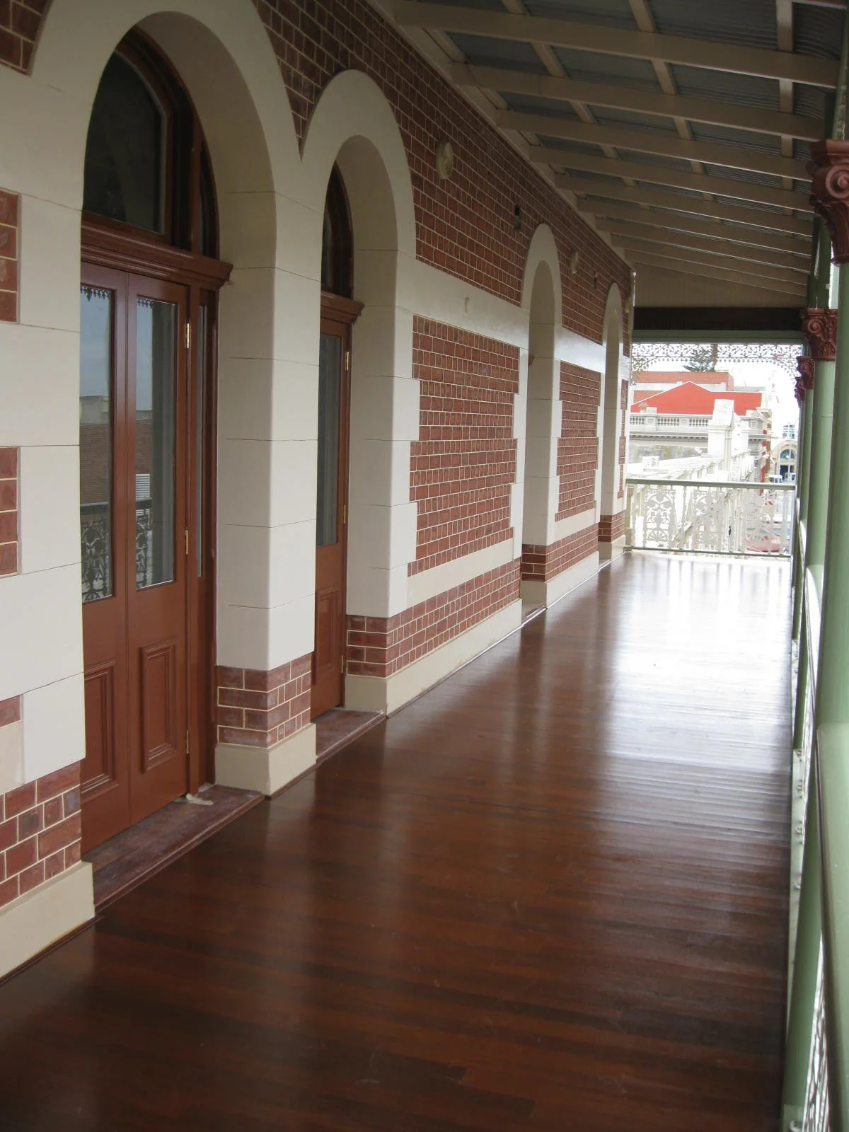 View of a balcony with hardwood flooring and a brick and white wall exterior, featuring arched windows and a railing, overlooking city buildings with red rooftops.