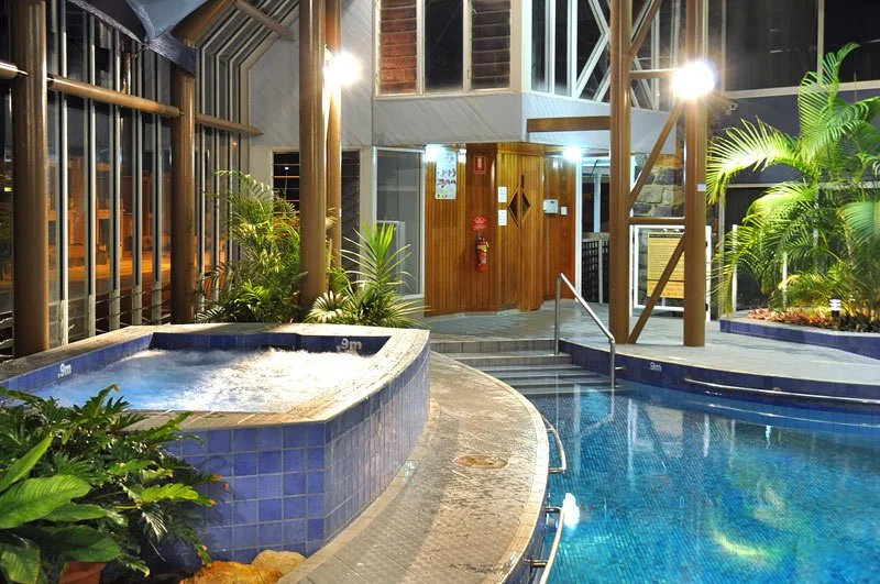 Indoor swimming pool and hot tub area with plants, glass walls, wooden accents, and lighting.