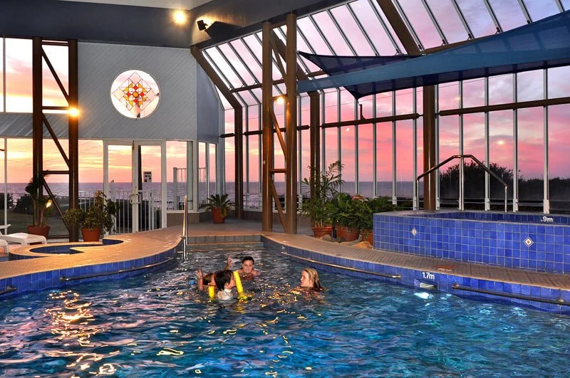 Indoor swimming pool with four people swimming and talking, large windows showing a sunset sky, potted plants, and a decorative stained glass window.