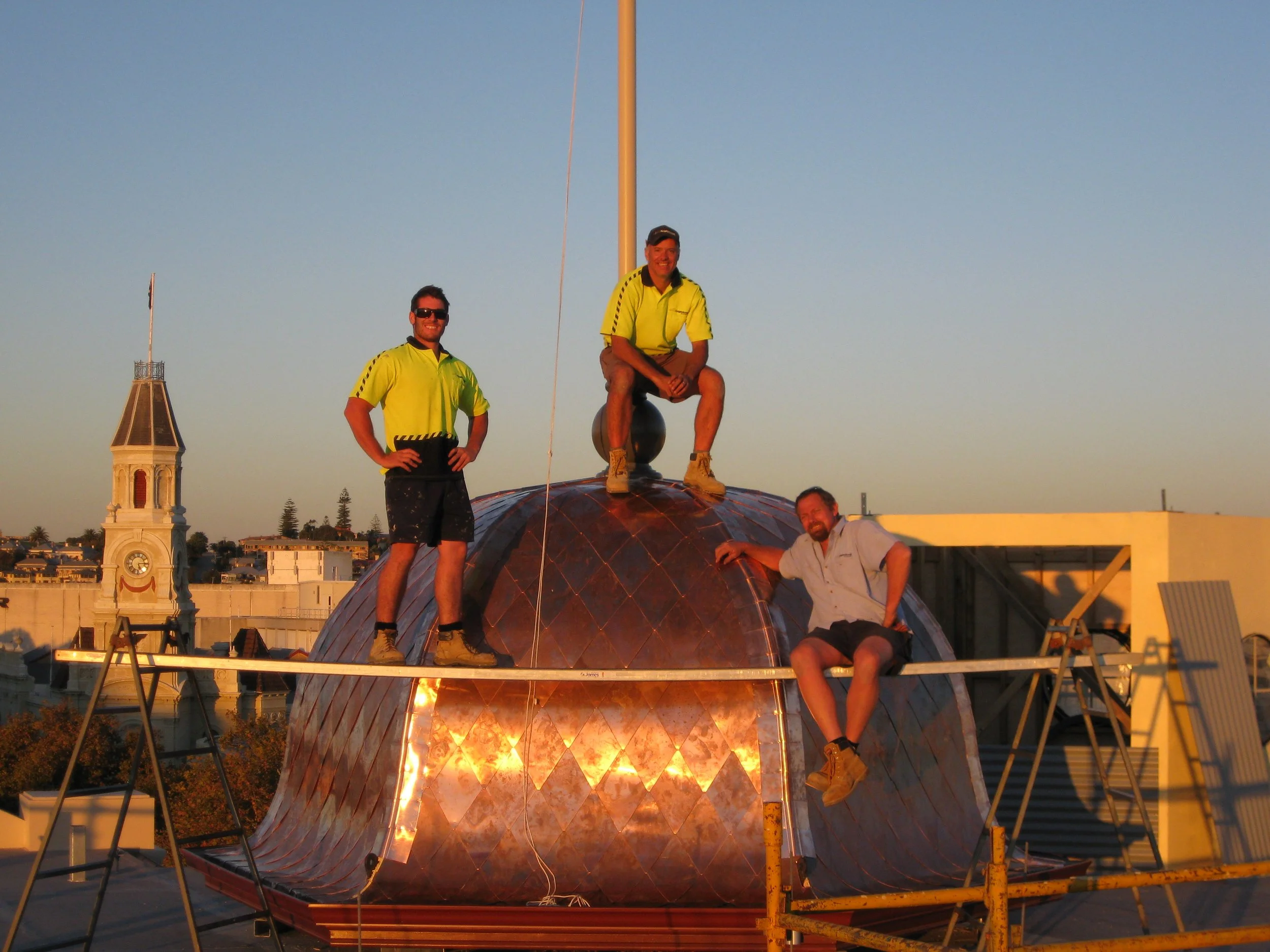 Four men working on a vaulted copper roof at sunset, with a clock tower and buildings in the background.
