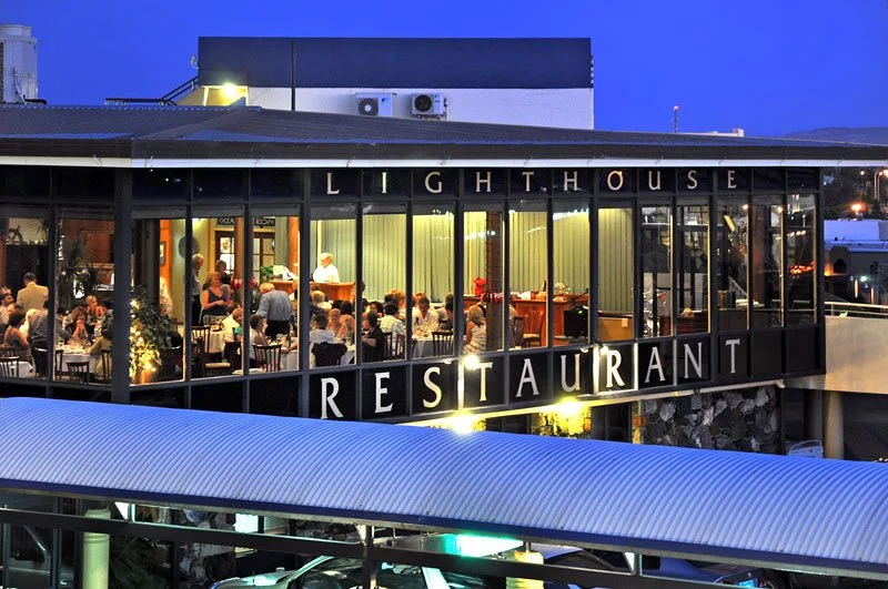 The Lighthouse Restaurant is a two-story building with large glass windows showing people dining inside during the evening. The restaurant's name is displayed with large black letters on the upper part of the building.