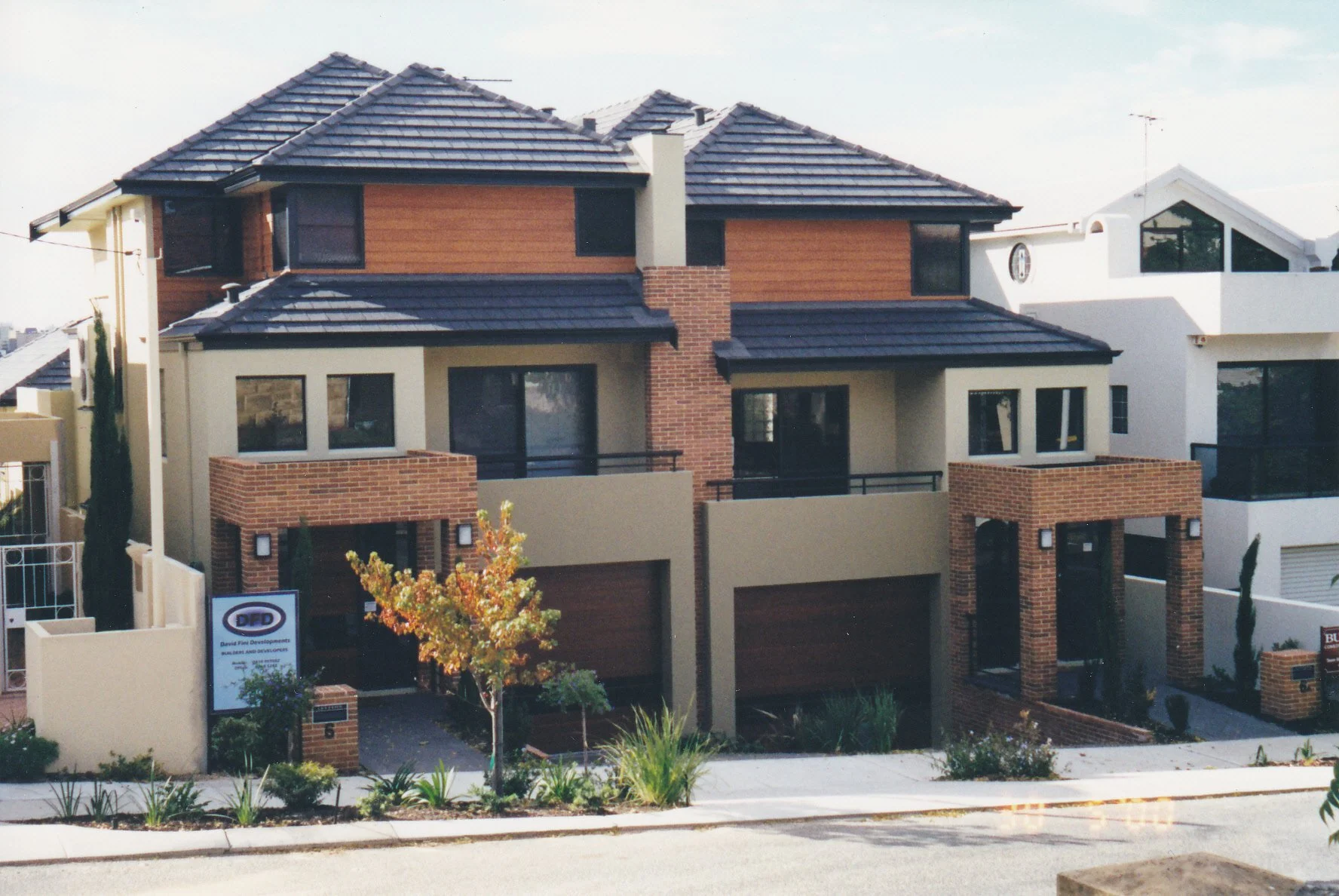 Multi-story residential building with brick and stucco exterior, tiled roofs, and front balconies, surrounded by a sidewalk and landscaping including small trees and shrubs.