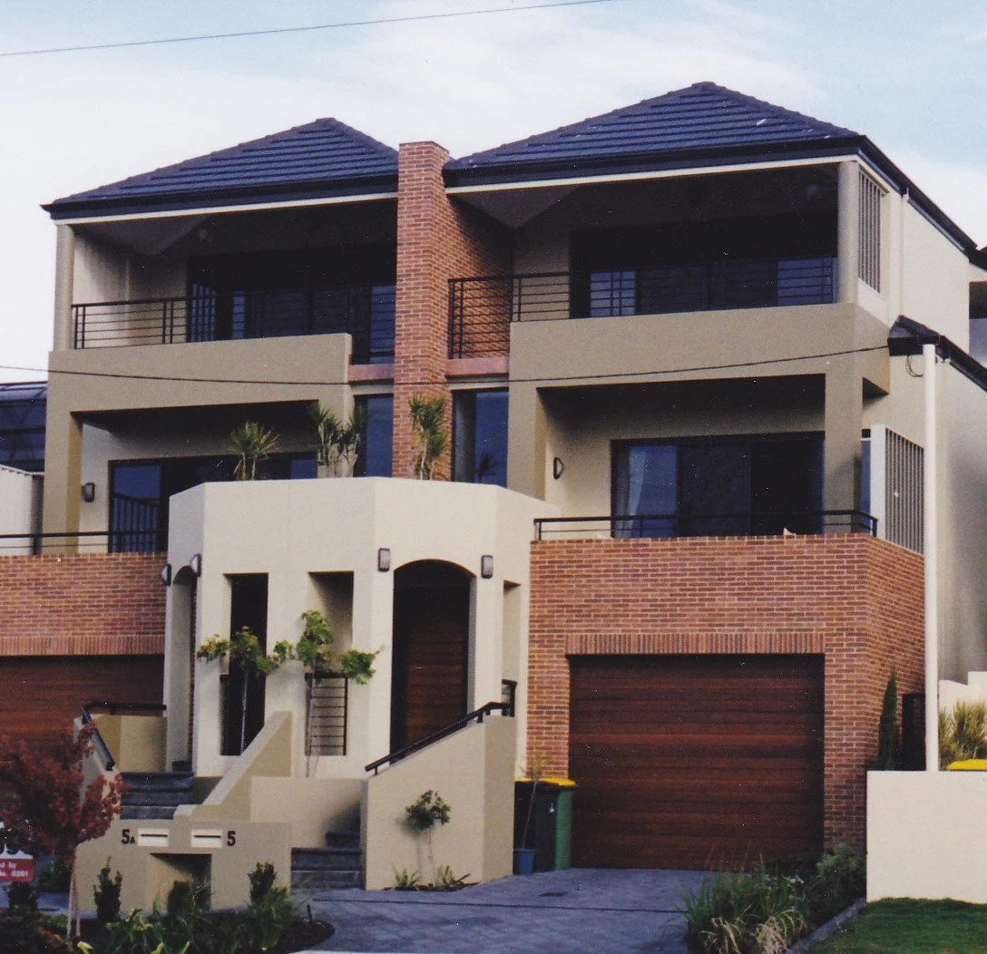 Modern multi-story residential house with beige and brick exterior, balconies, and a garage. Landscaping includes trees and plants.