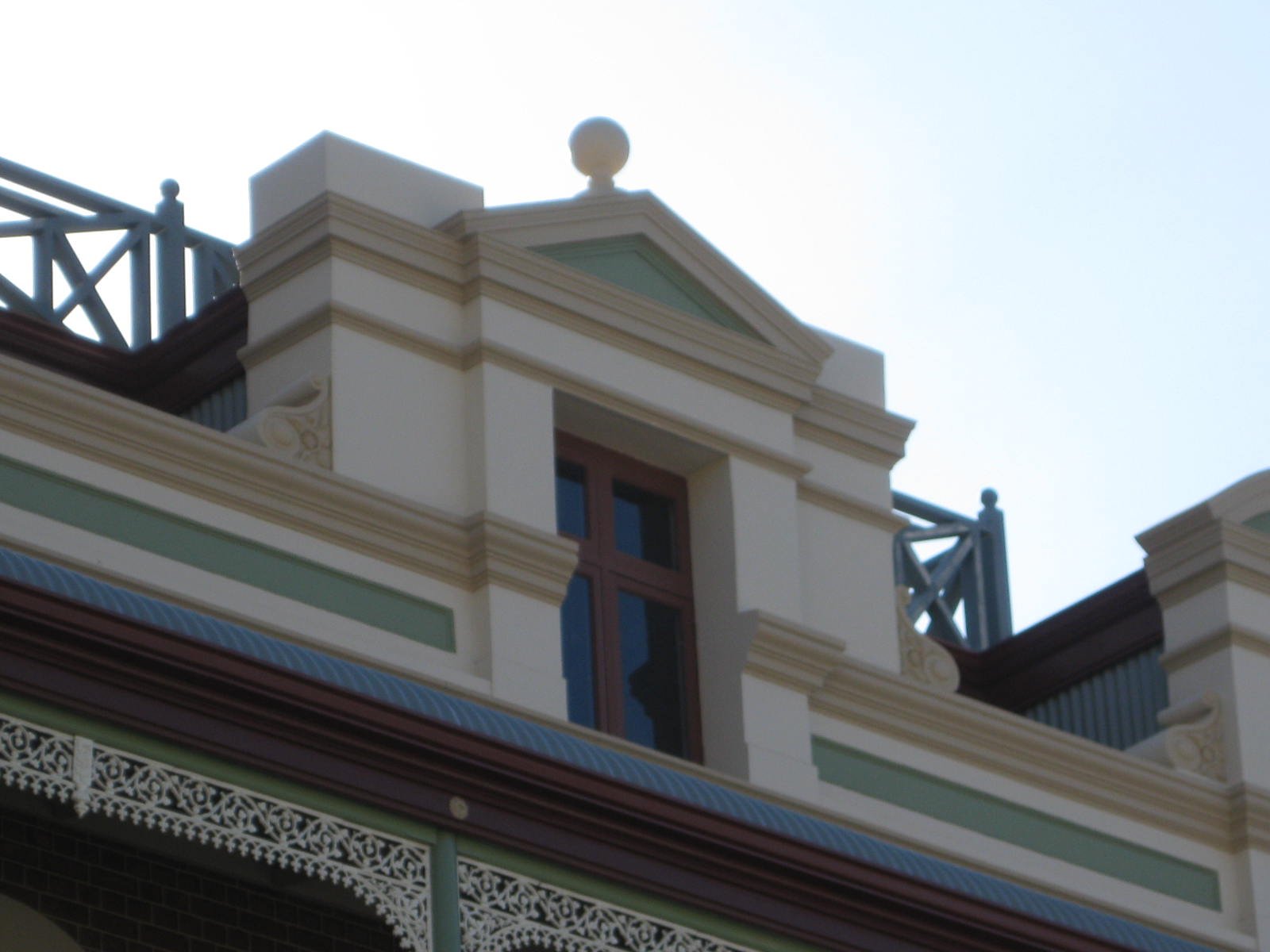 Close-up of the upper part of a historic building with decorative trim, ornate moldings, and a small balcony with turnings, beige and green color scheme, and modern railings on the roof.