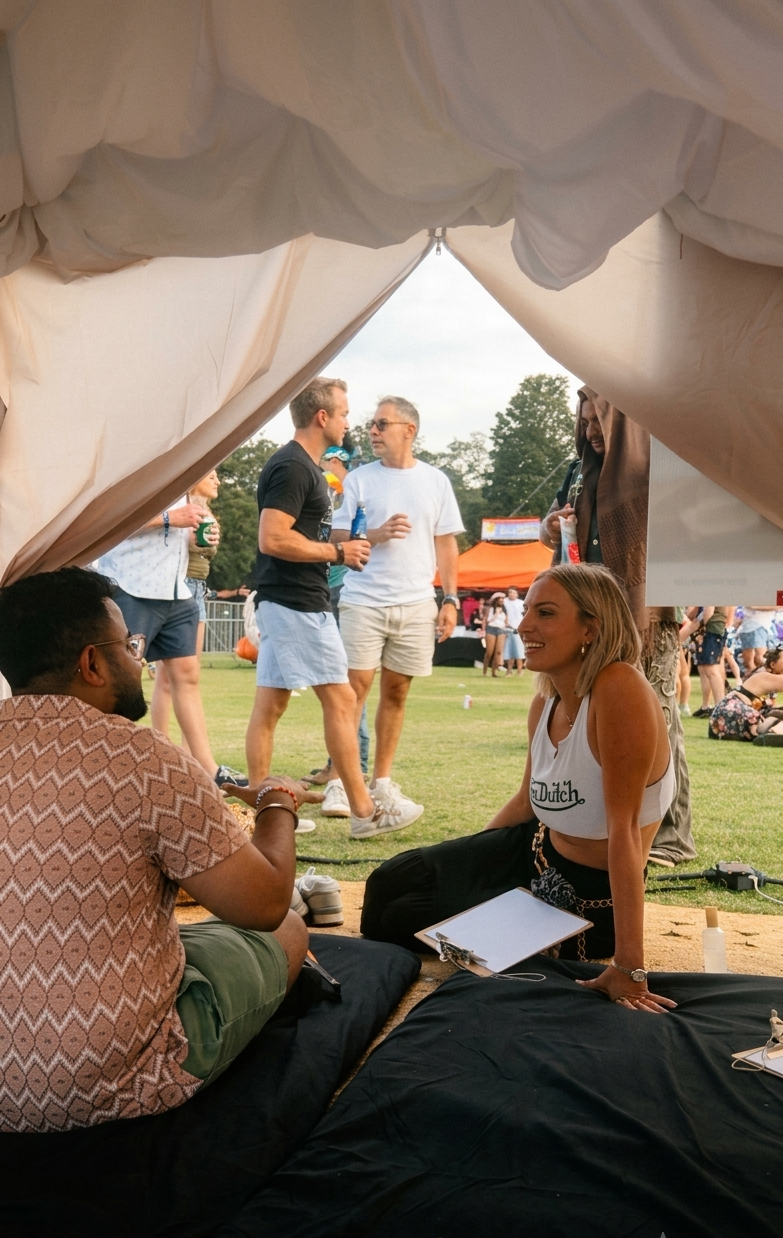 People socializing at an outdoor event, with some sitting under a tent and others standing in the background, on a grassy area during daytime.