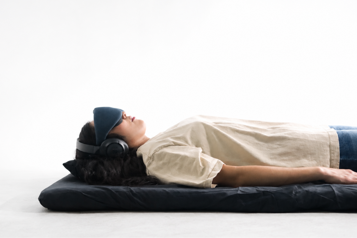A woman lying on a black mat on the floor, wearing headphones, an eye mask, a beige t-shirt, and blue jeans, resting with her eyes closed.