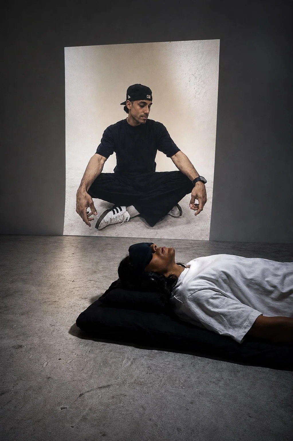 A man sitting in a cross-legged position on the floor, seen through a square mirror on the wall. A woman is lying on the floor with a pillow and headphones, wearing a sleep mask and white shirt.