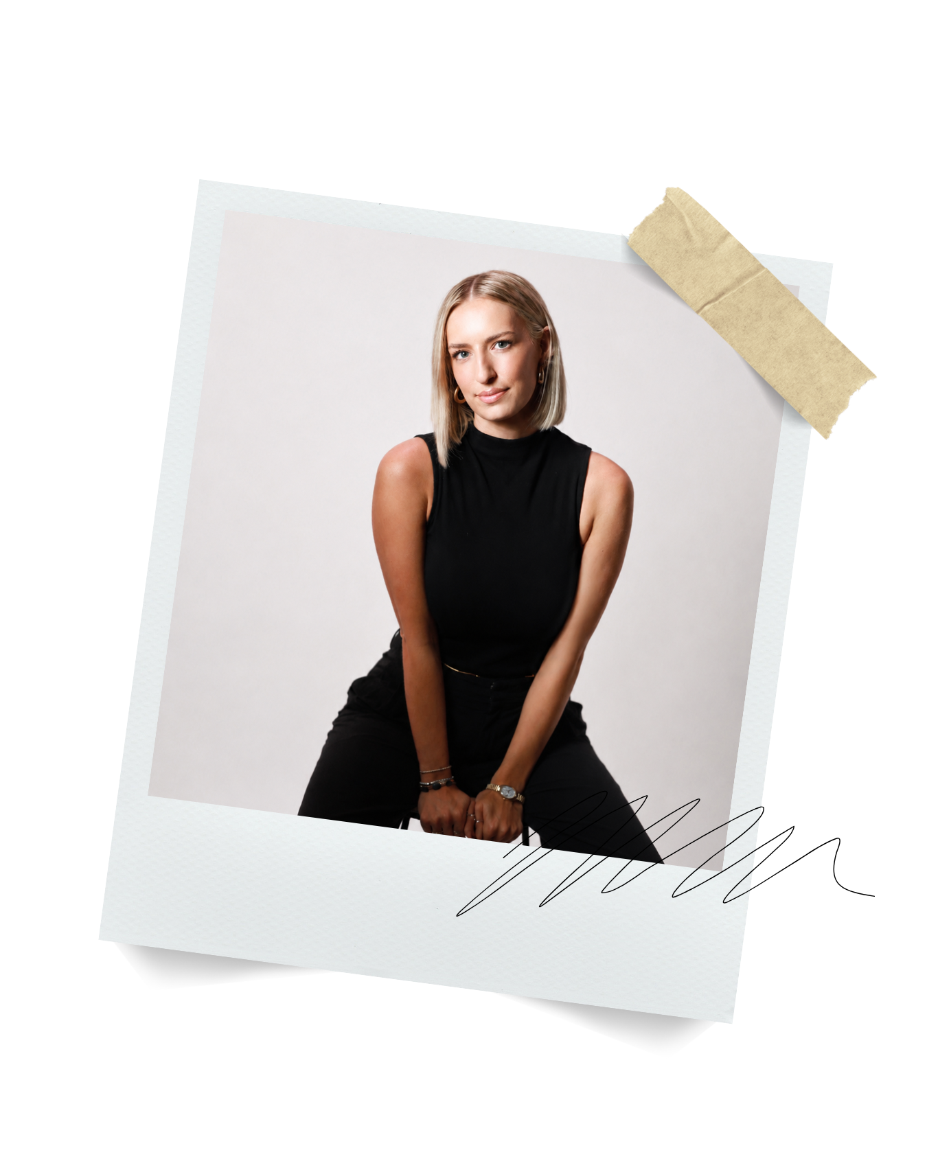 A woman with blonde hair and light skin sitting against a plain white background. She is wearing a sleeveless black top, dark pants, and jewelry, looking at the camera.