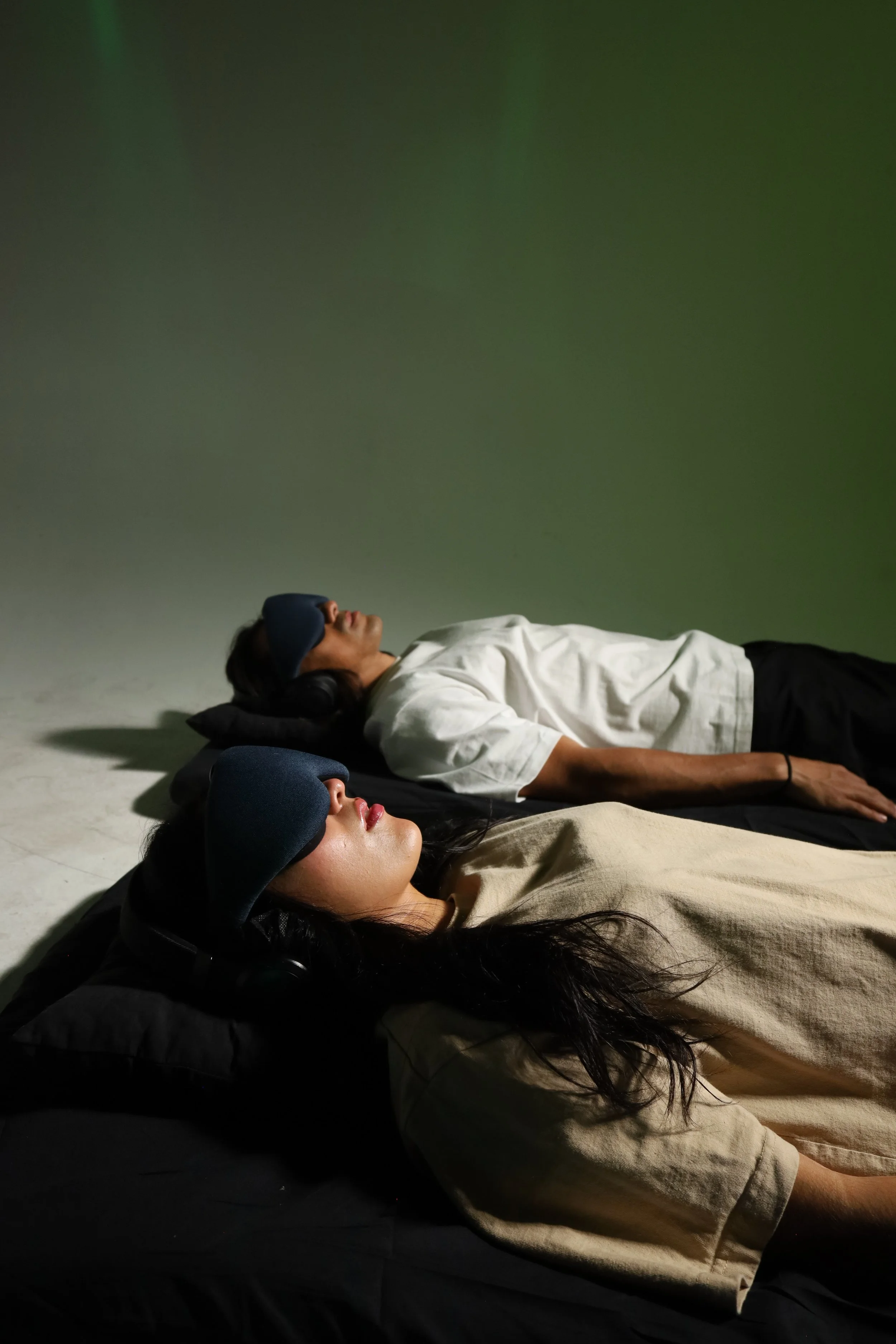 Two people lying on their backs on the floor in a room with a plain green wall. They are wearing sleep masks and headphones, appearing to relax or rest. One individual has long dark hair and the other has shorter dark hair.