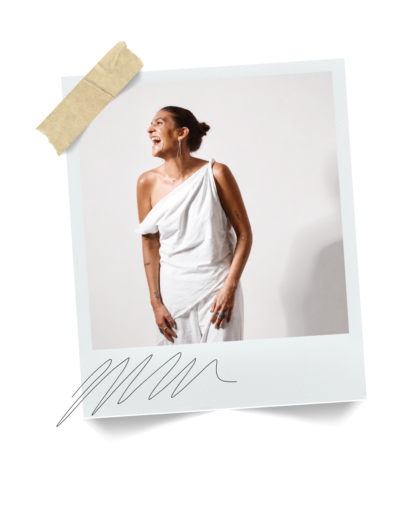 A woman with tattoos on her arms, wearing a white, one-shoulder dress, laughing and standing against a white background.