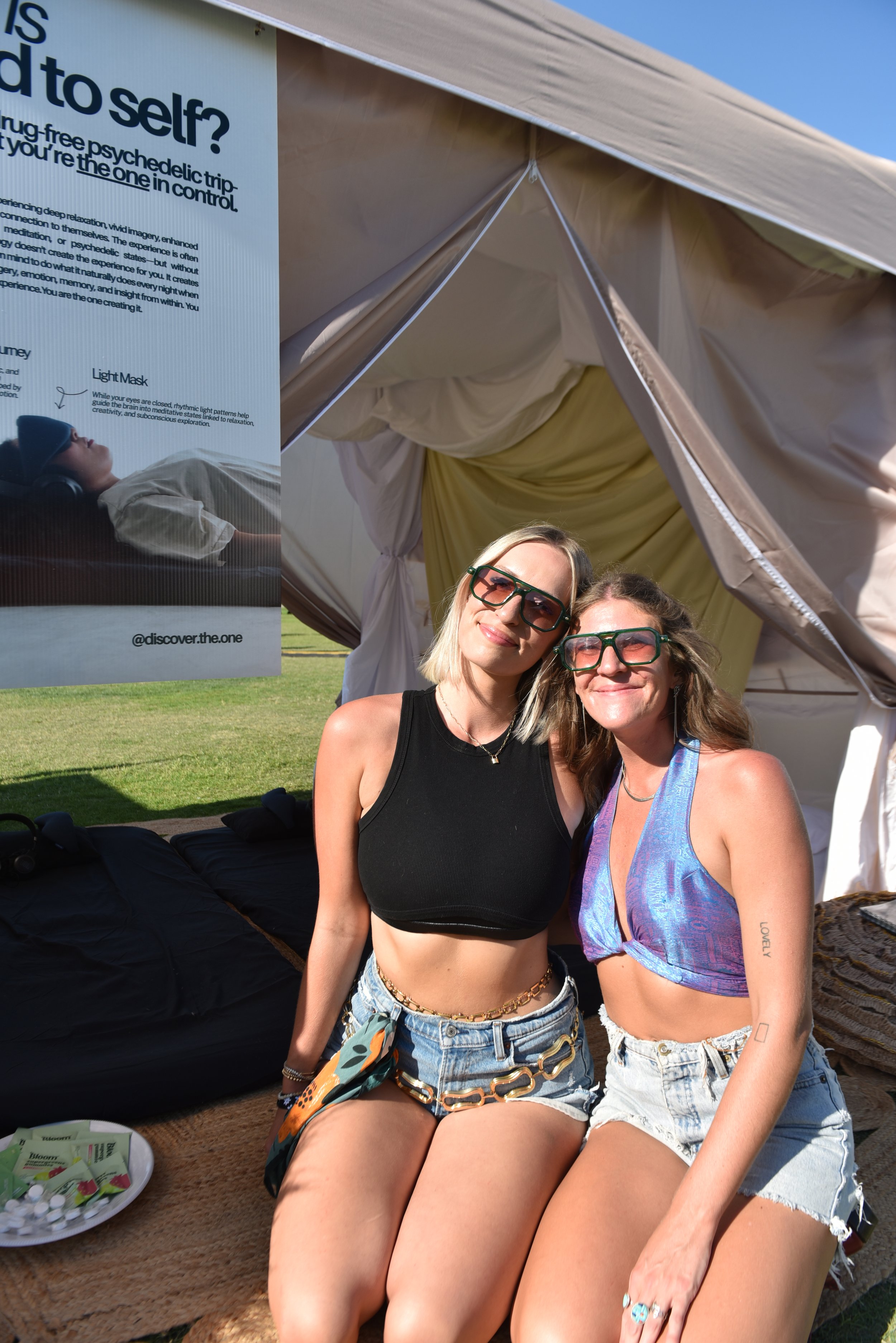 Two women sitting outdoors under a tent, wearing sunglasses and summer clothing, smiling at the camera. A poster about psychedelic trips and a tray of supplies are nearby.