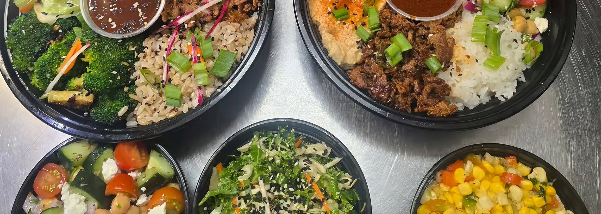 Assorted bowls of salads and rice dishes with vegetables, greens, cherry tomatoes, corn, and beef, served in black containers.
