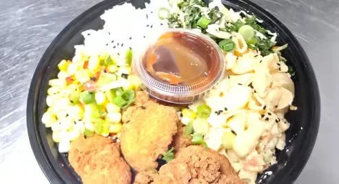 A bowl with white rice, chopped green onions, shredded chicken, fried chicken pieces, and a small container of sauce.