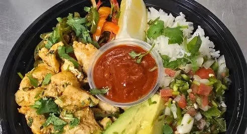 A black takeout container with grilled chicken, mixed vegetables, white rice with green herbs, avocado slices, lemon wedge, and a small cup of red sauce.