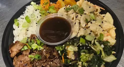 Plate of food with white rice, grilled meat, macaroni salad, greens, a small container of dark sauce, and some vegetable sides