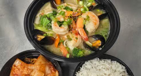 Three black takeout containers, one with creamy shrimp and vegetable soup with shrimp, broccoli, carrots, onions, and green onions, another with cooked white rice, and a third with salmon fillet.
