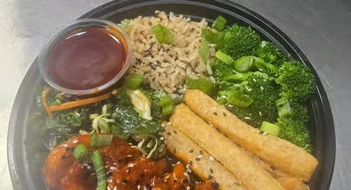 A black takeout container filled with rice, broccoli, mixed vegetables, two spring rolls, a piece of fried chicken, and a small container of dipping sauce.