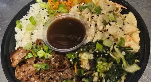 Plate with white rice, grilled beef, cooked vegetables, chopped green onions, and a small cup of soy sauce.