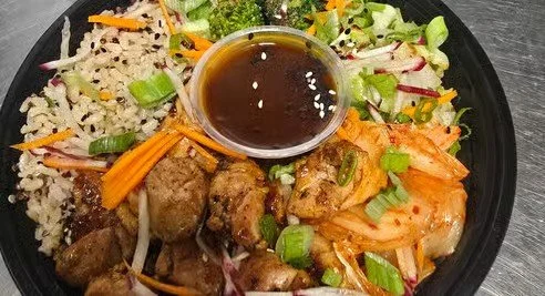 A bowl of Asian-inspired cuisine with grilled meat, fresh salad greens, shredded vegetables, kimchi, and a small dish of soy sauce or dressing.