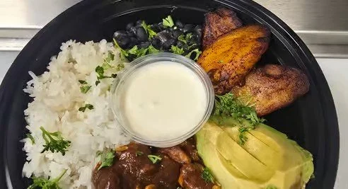 A plate with white rice, black beans, grilled plantains, avocado slices, a small cup of white sauce, and beef stew topped with green herbs.