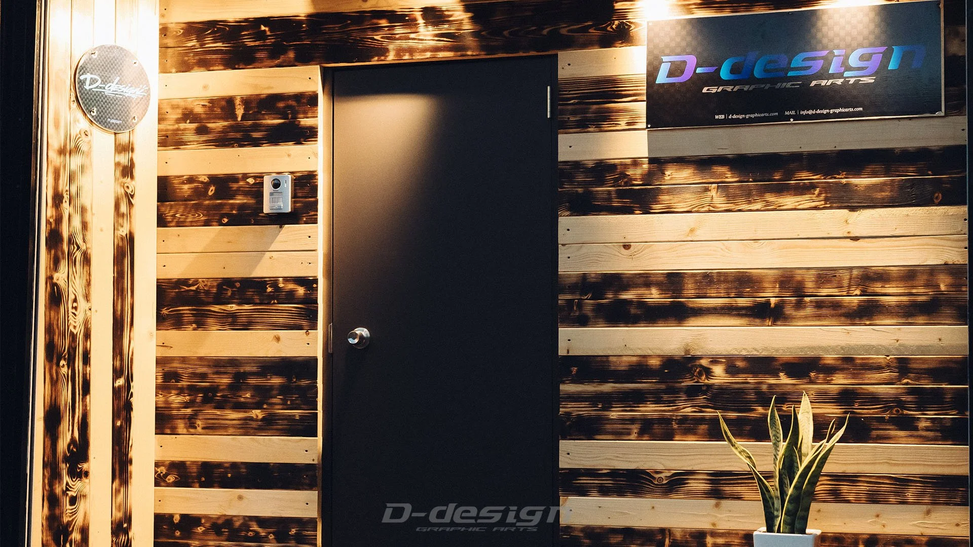 木の板張りの壁に黒いドアとD-design GRAPHIC ARTSの看板が設置されている正面玄関の風景。