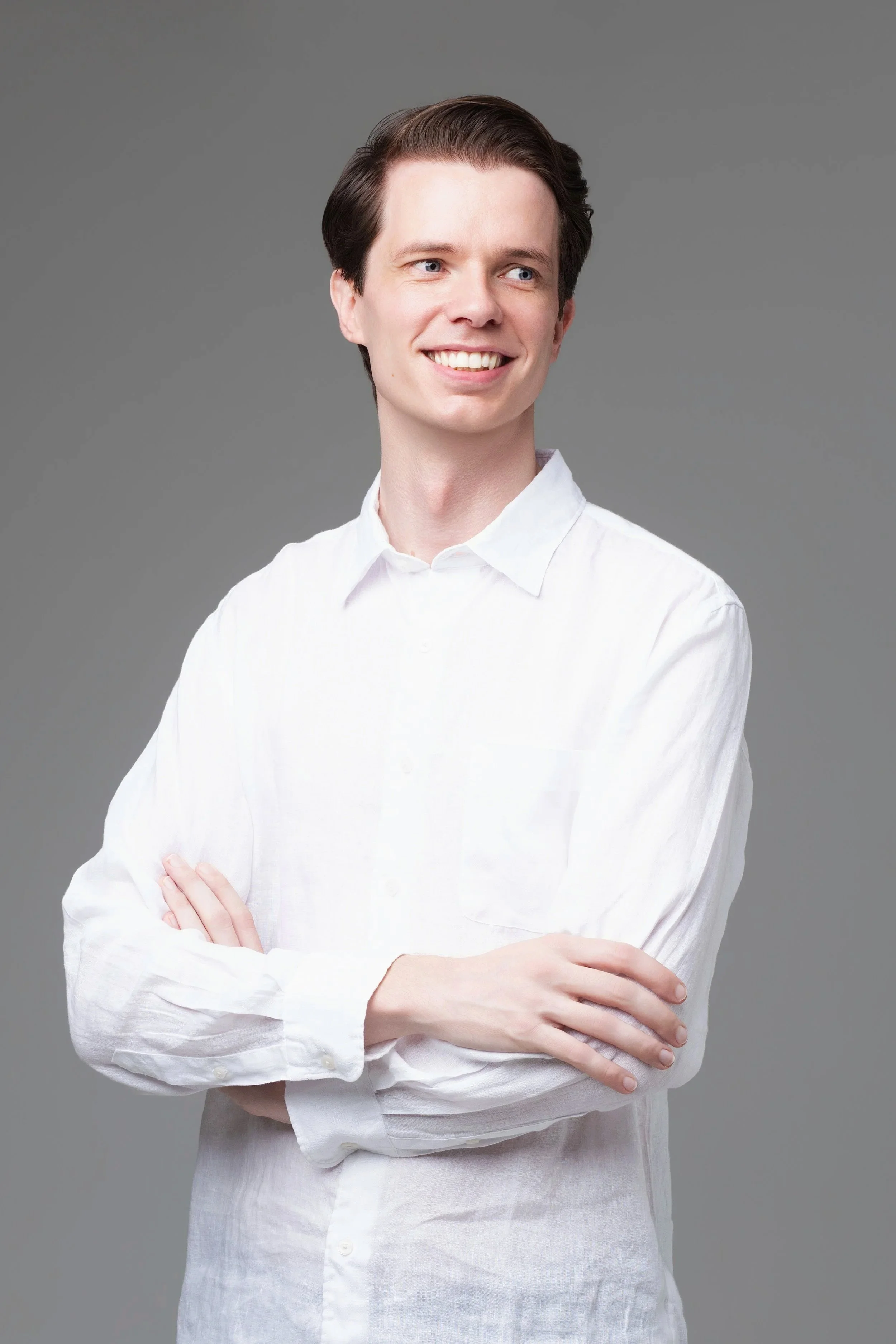 Headshot of a young man with dark hair, wearing a white shirt, smiling, with arms crossed against a gray background.