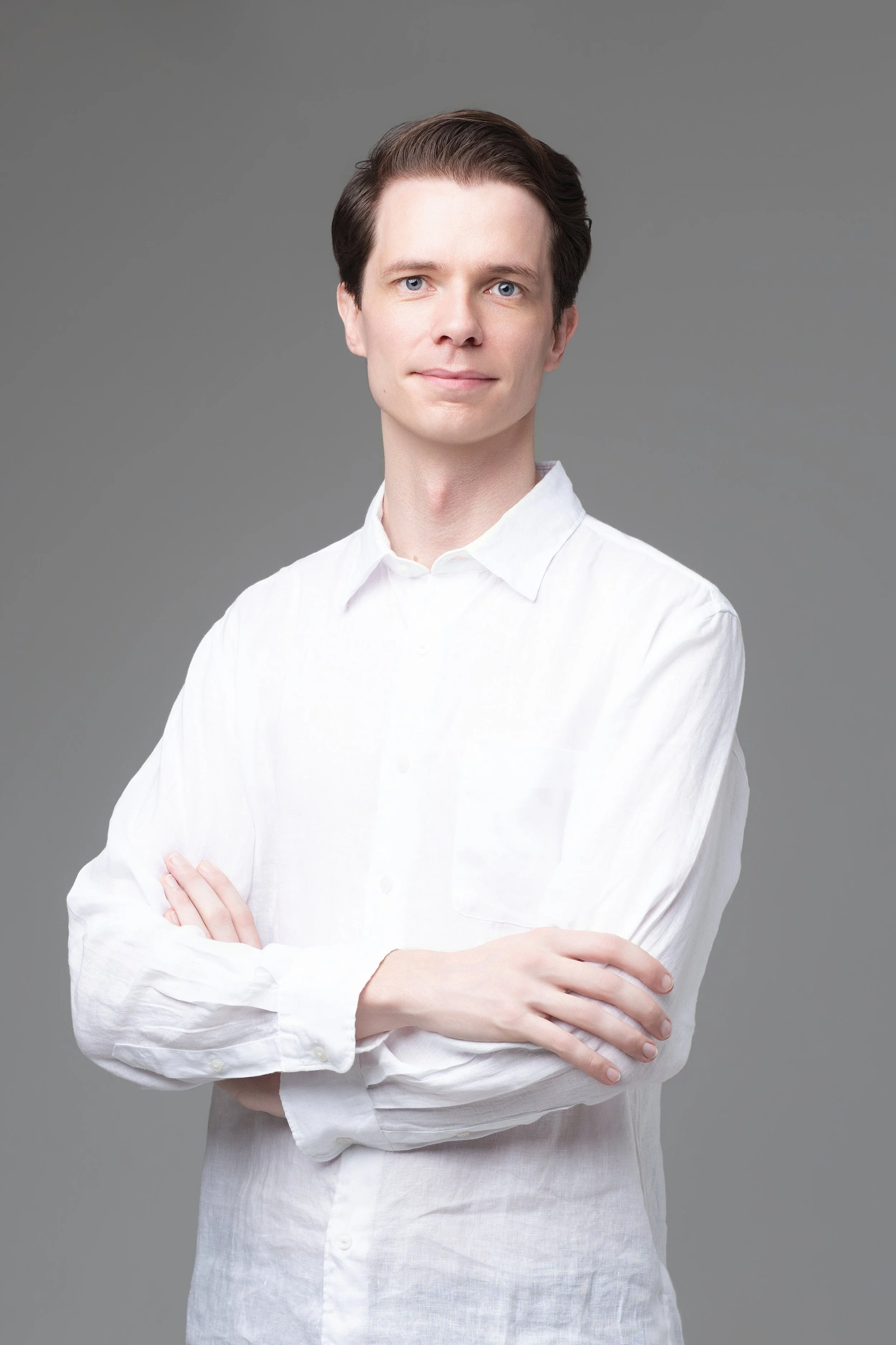 A young man with blue eyes and dark brown hair styled back, wearing a white button-up shirt, standing against a neutral gray background, with arms crossed and a slight smile.
