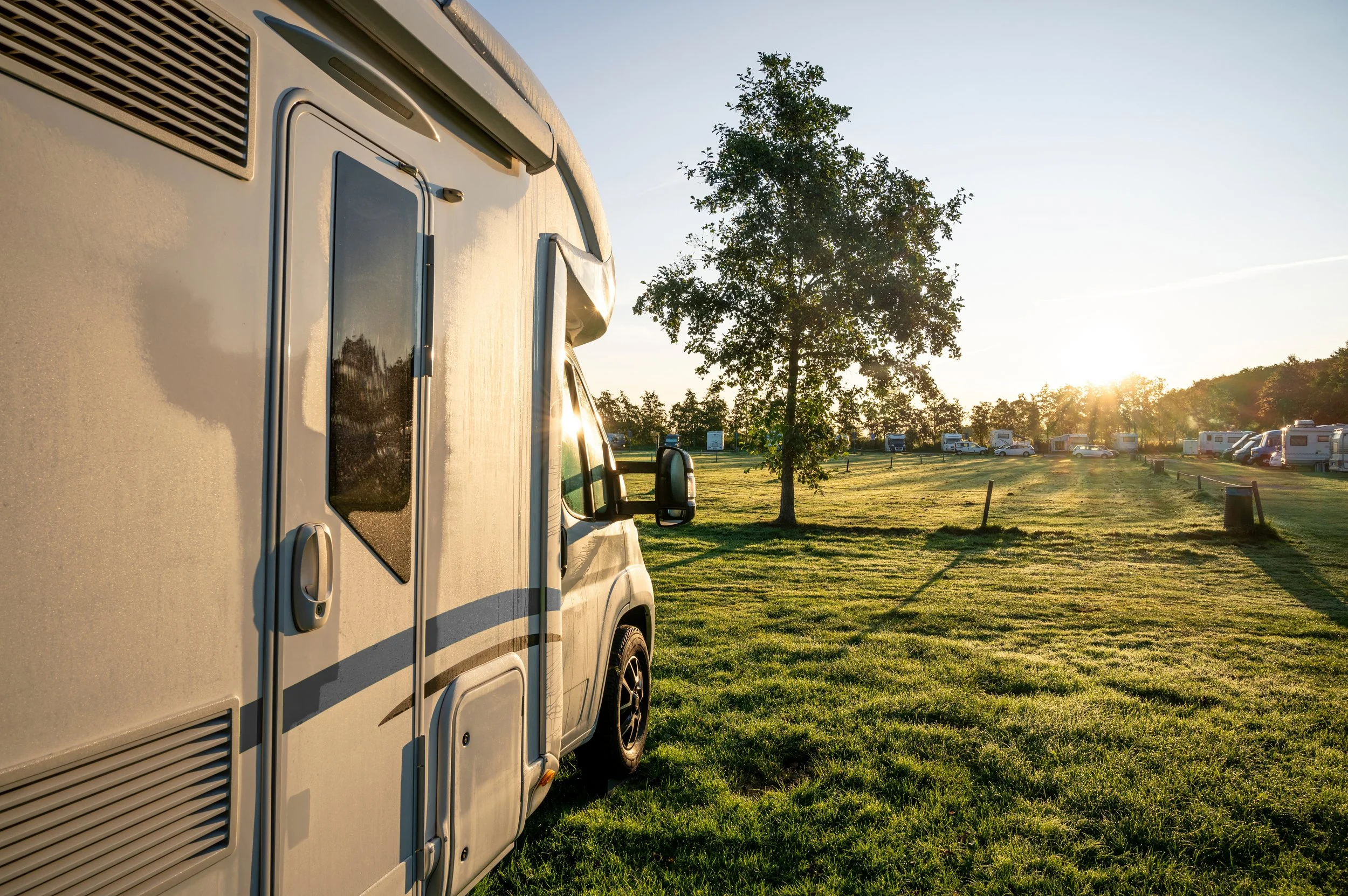 A white camper van parked on a grassy field during sunset, with trees and other RVs in the background.