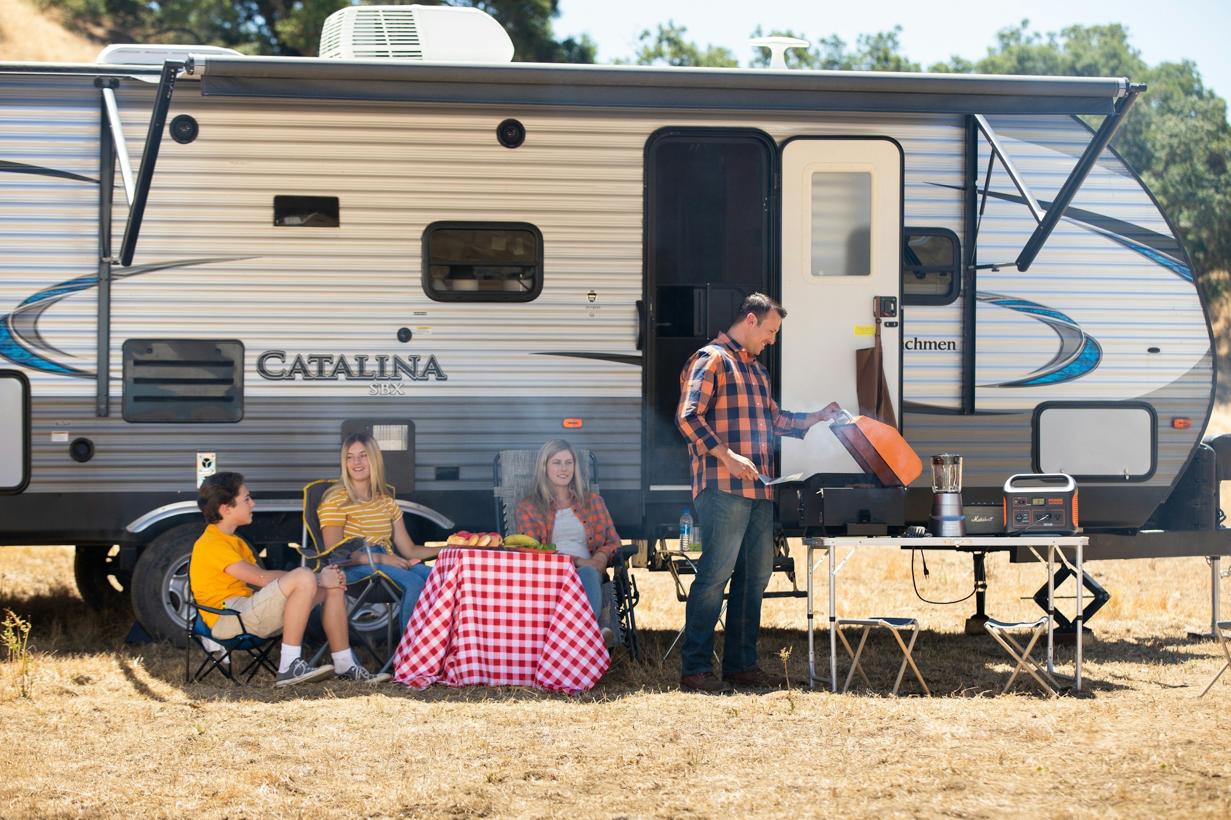 A group of people having a picnic outside a travel trailer. One man is grilling food, and three women and a boy are sitting in chairs around a table with a red and white checkered tablecloth. The scene is in a grassy area with trees in the background.