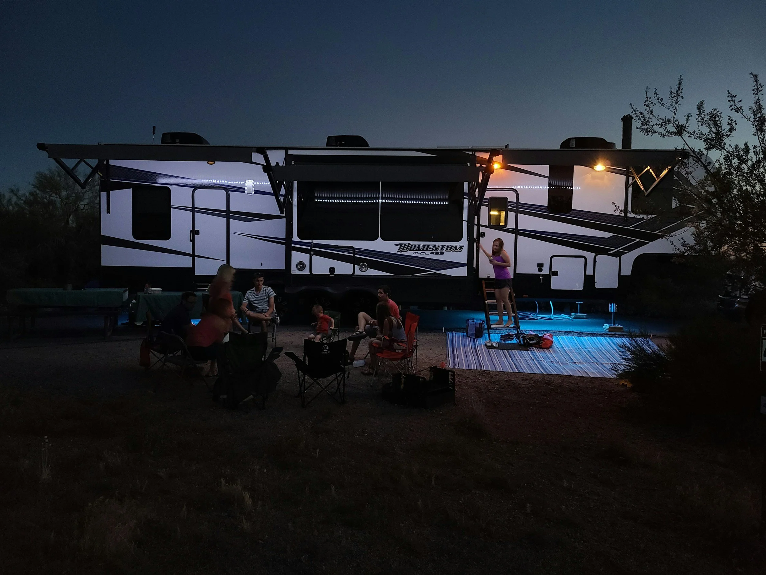 A large white and black recreational vehicle (RV) parked outdoors at dusk, with a group of people sitting and standing around, and a woman in a purple shirt standing on a step ladder. There are trees and outdoor lighting in the scene.