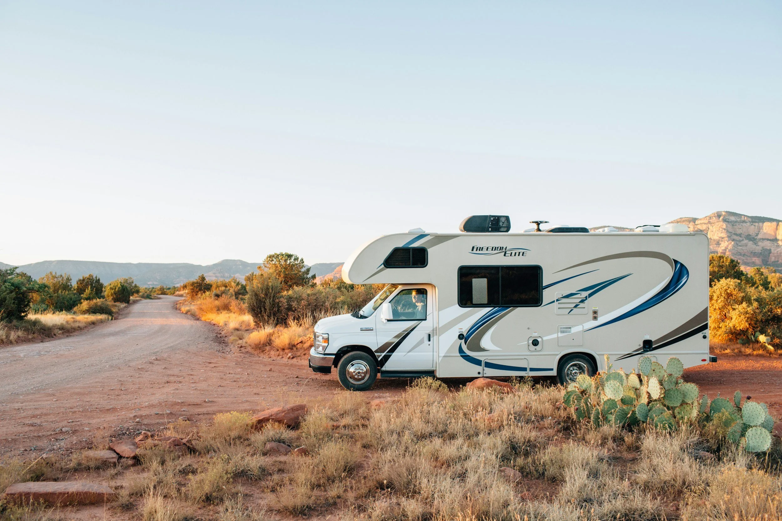 A white recreational vehicle with blue and gray graphics parked along a dirt road in a desert landscape with sparse vegetation, cacti, and mountains in the background during sunset.