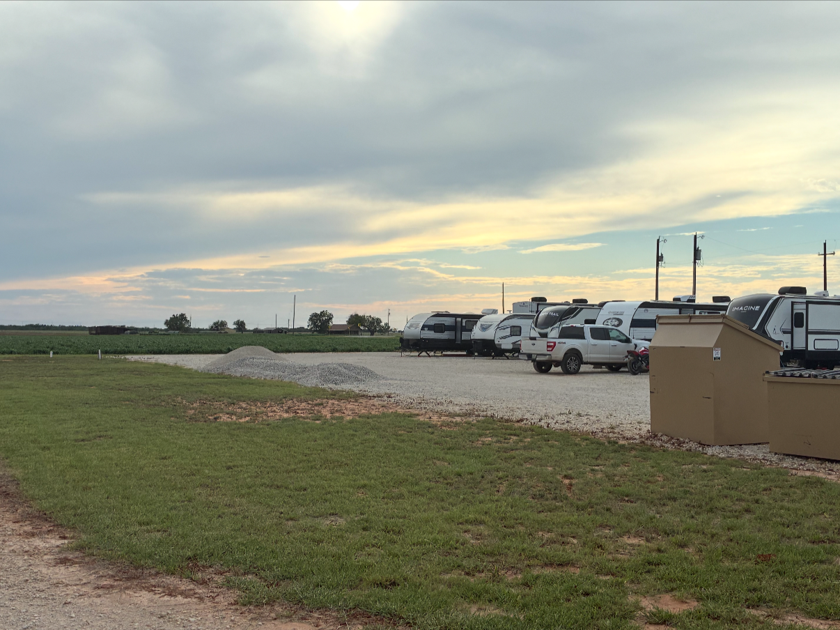 A parking lot with several RVs and a pickup truck, next to grassy and gravel areas under a cloudy sky at dusk or dawn.