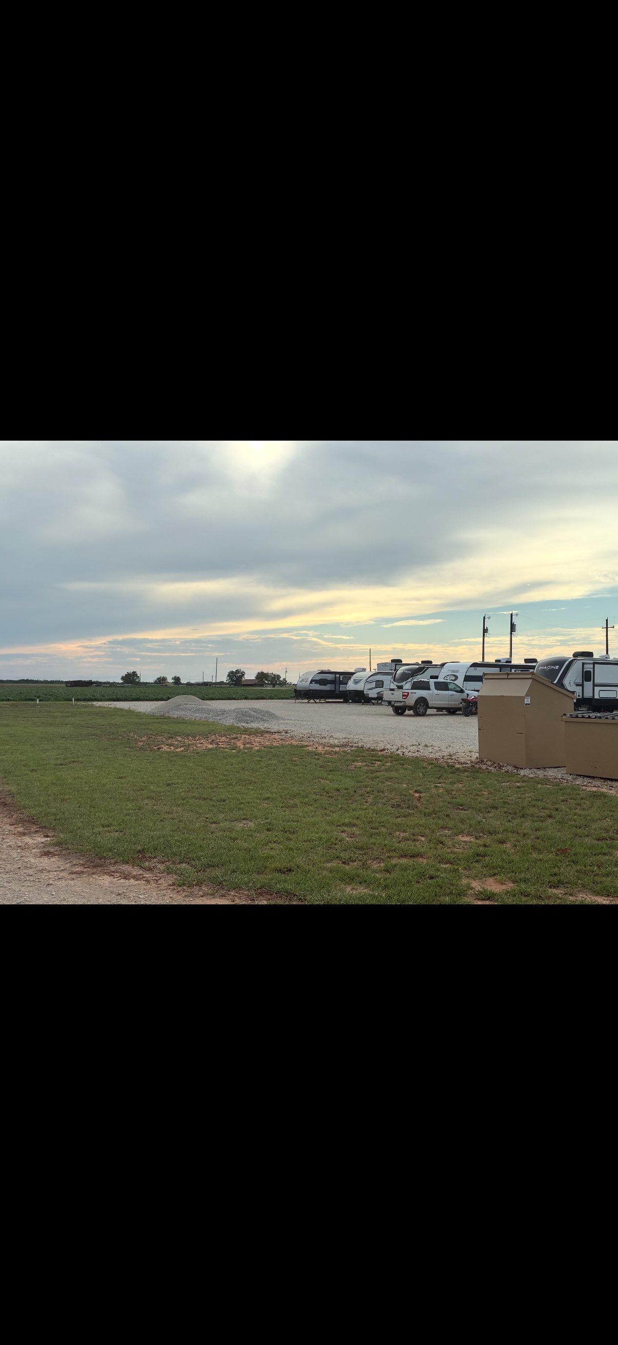 A parking lot with several RVs, travel trailers, and cars under a cloudy sky with the sun setting.