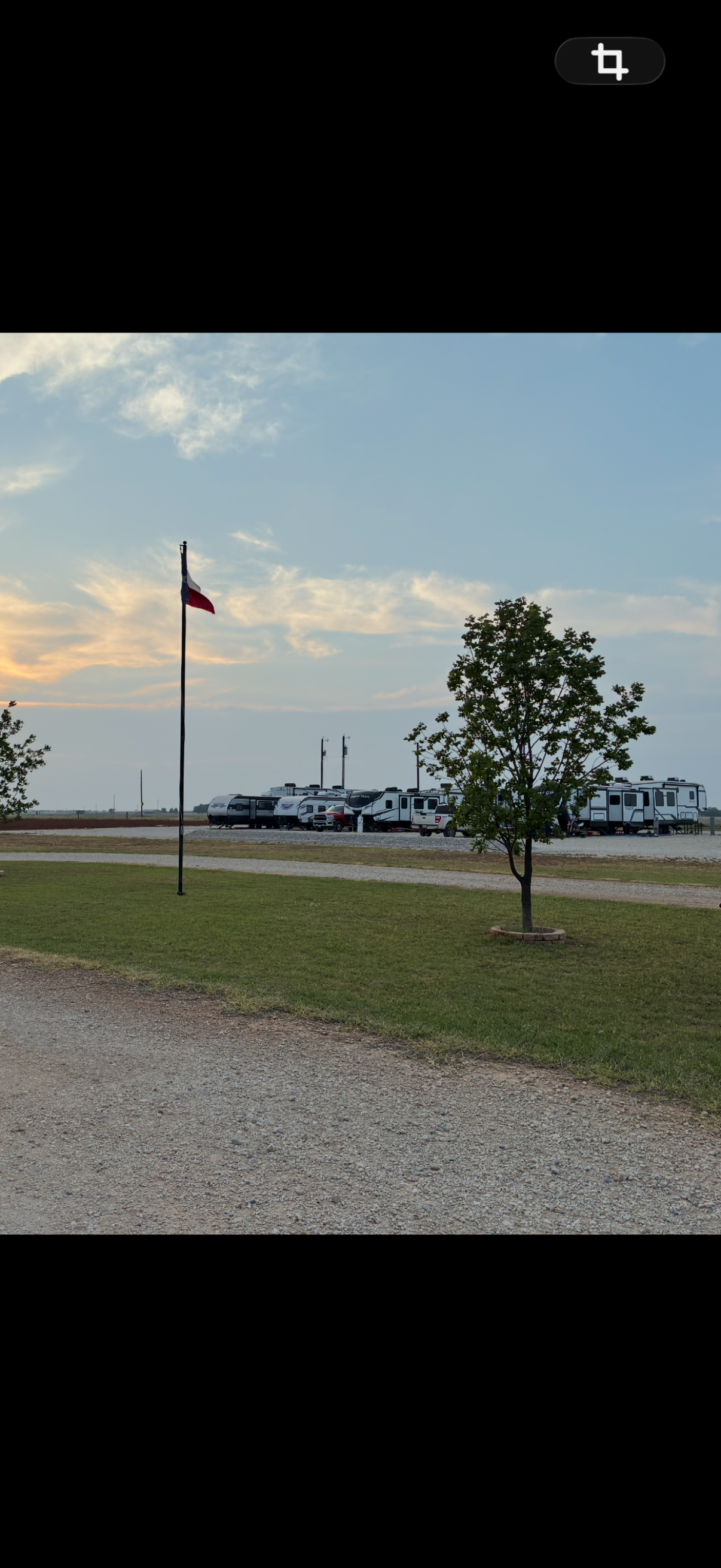 A flagpole with the Texas state flag, several RVs and trailers parked in the background, a tree in the foreground, and a sunset sky with clouds.