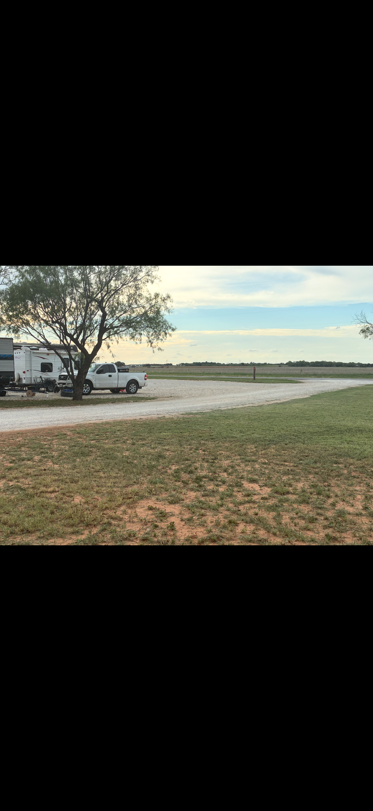 A rural scene with a gravel road, a tree, and a white pickup truck parked beside a camper trailer, with an open field and partly cloudy sky in the background.