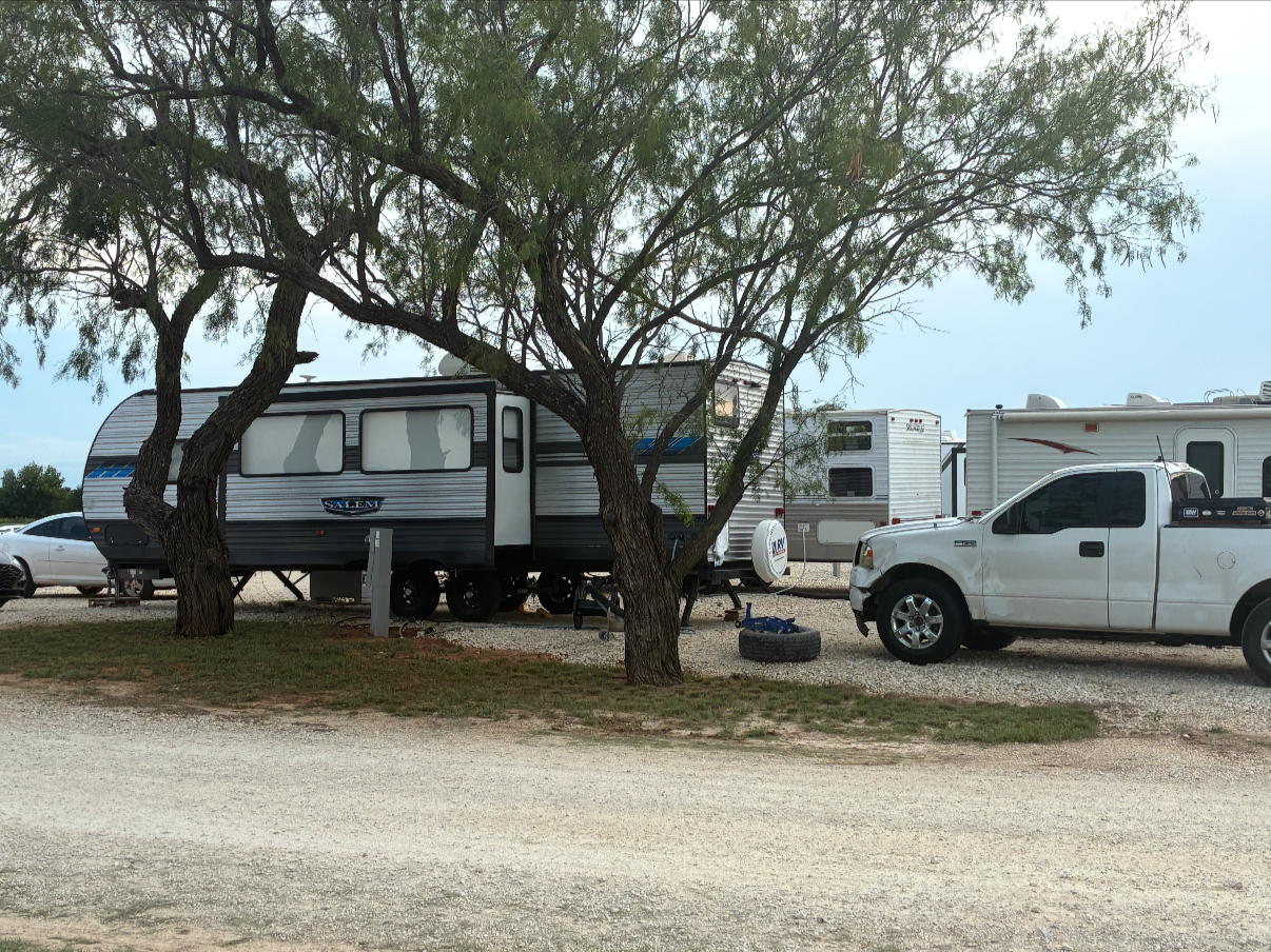 A recreational vehicle park with a trailer, a pickup truck, and trees on a gravel lot under a partly cloudy sky.