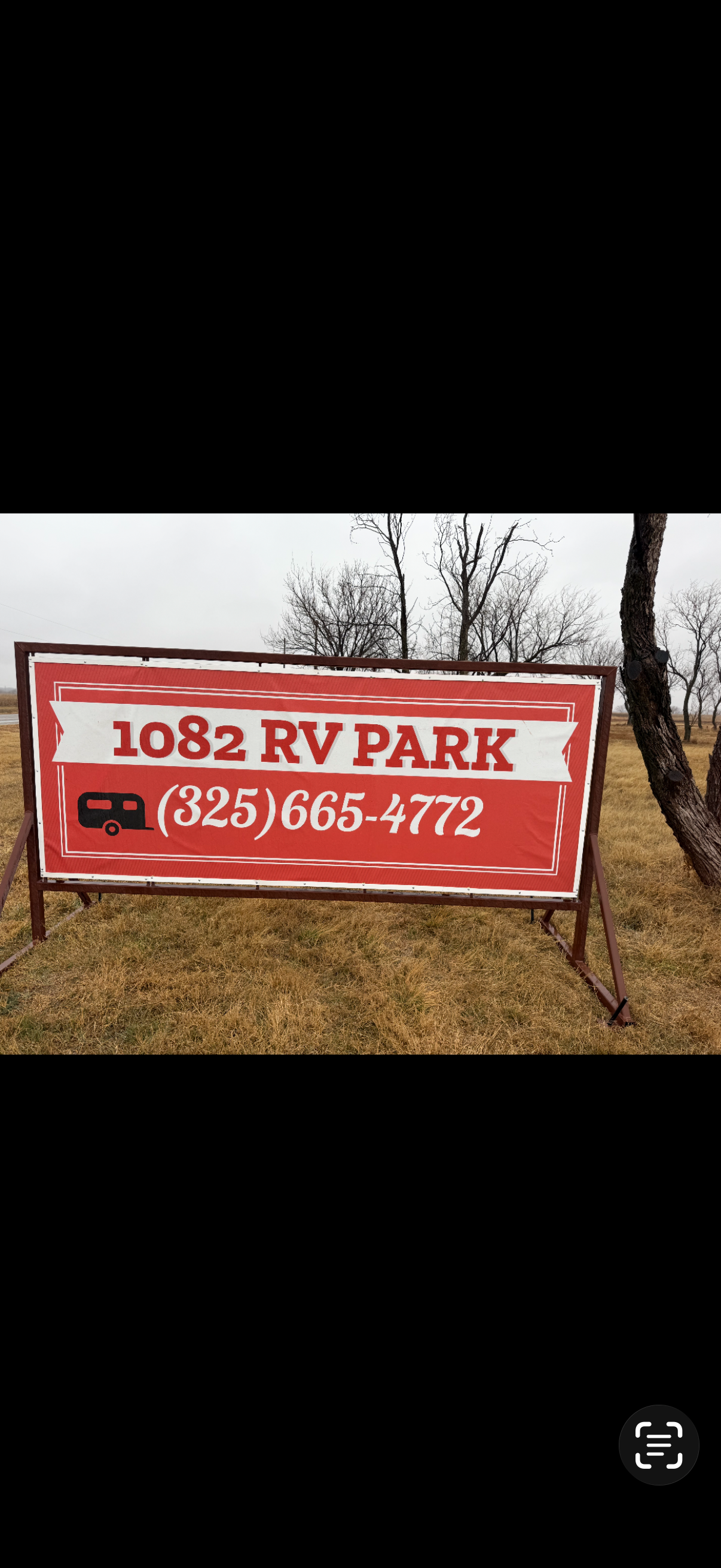 Red and white sign for 1082 RV Park, displaying a phone number (325) 665-4772, with a black RV icon, outdoors with leafless trees and cloudy sky in the background.