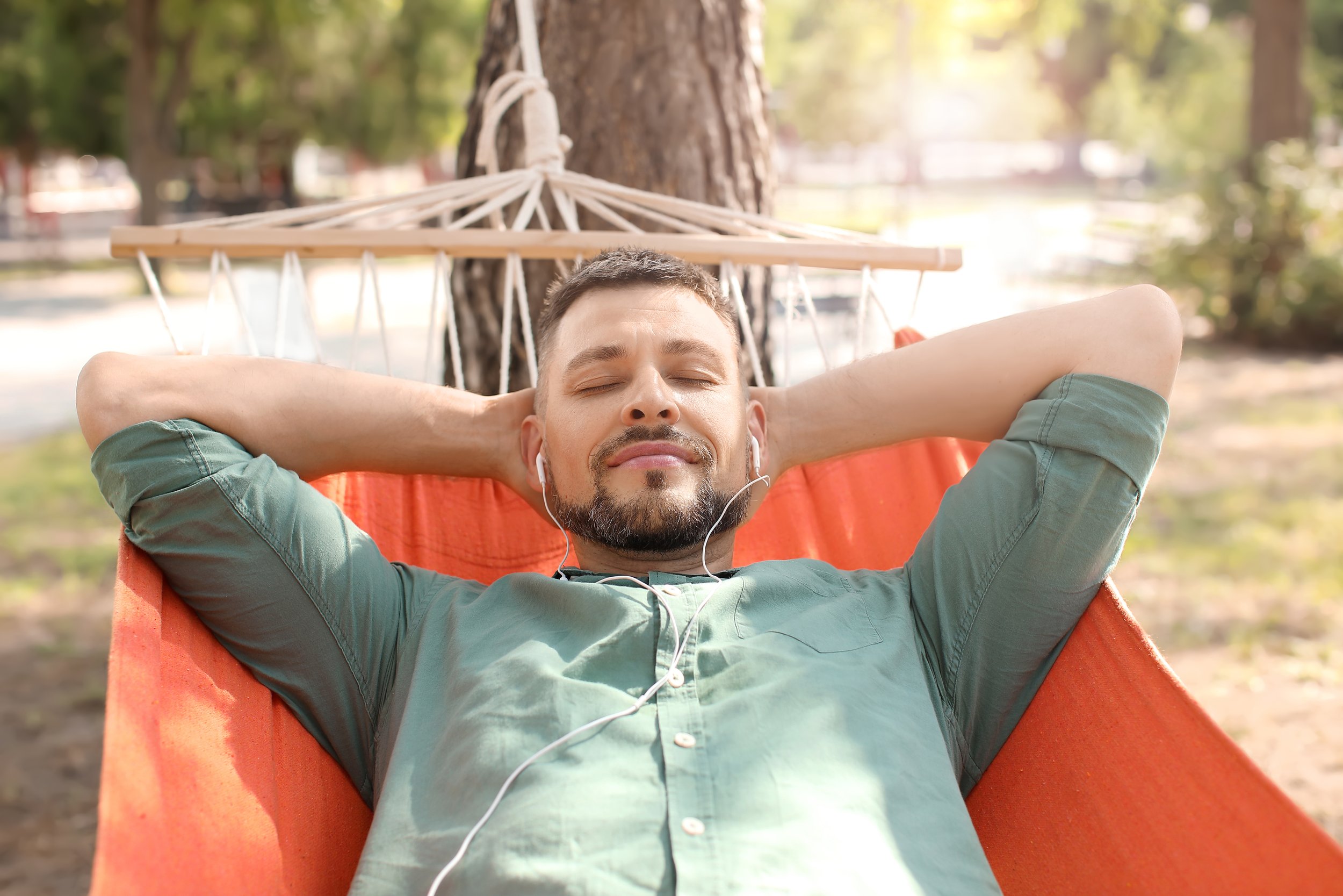 A man relaxing in an orange hammock tied to a tree in an outdoor park, listening to music with earphones and with eyes closed.