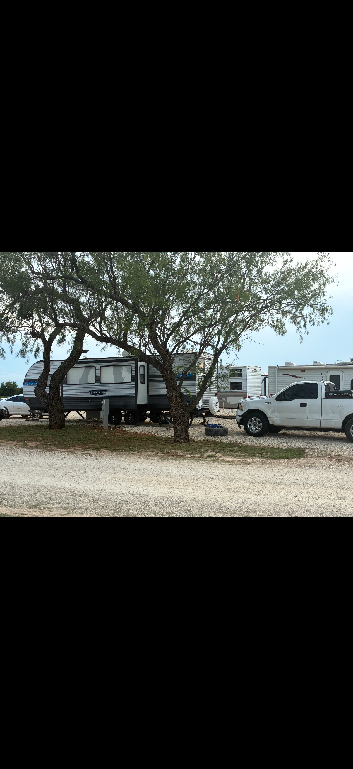 A row of RV trailers and a white pickup truck parked under trees at a campground, with a gravel ground and a cloudy sky.