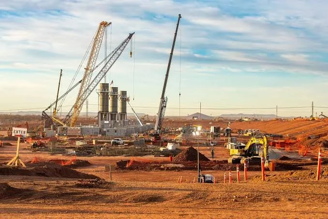 Construction site with cranes, construction vehicles, and large industrial equipment in a desert landscape at sunset.
