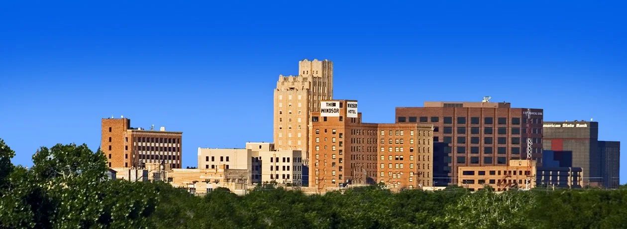 Panoramic view of a city skyline with various tall buildings, trees in the foreground, and a clear blue sky.
